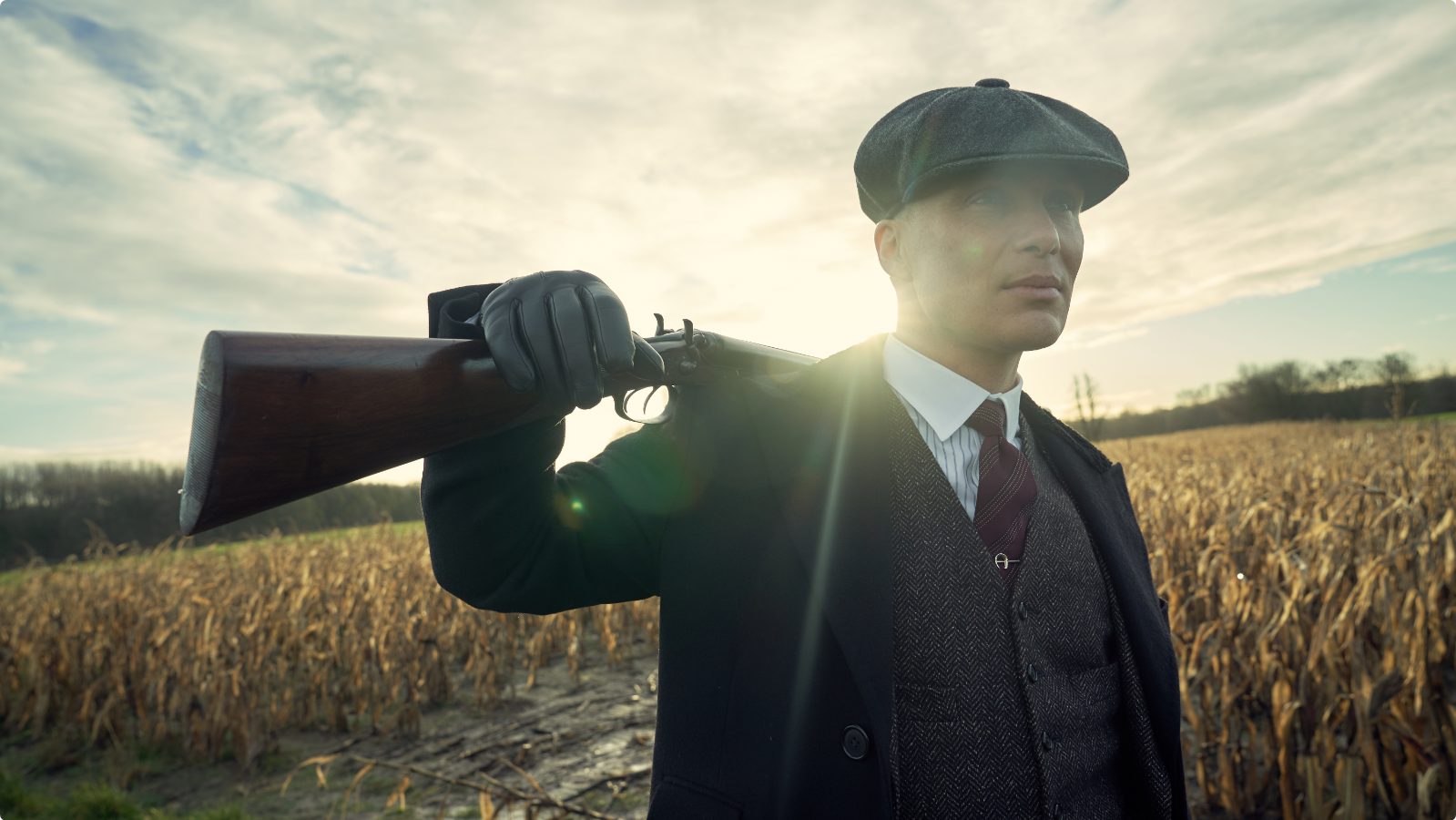 A man in vintage attire, including a flat cap, holds a shotgun over his shoulder, standing in a cornfield under a bright sky.