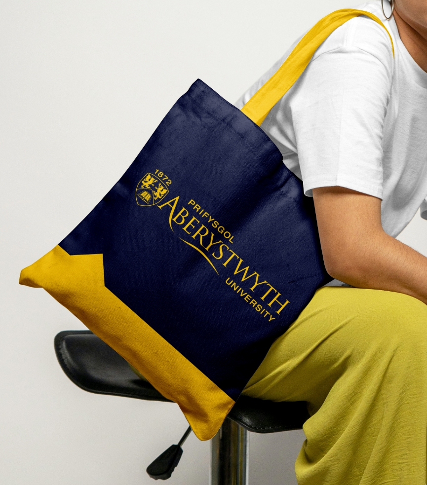 A person seated on a stool, partially visible wearing a white shirt and yellow pants, holds a navy and gold Aberystwyth University tote bag designed by a Brand Agency UK.