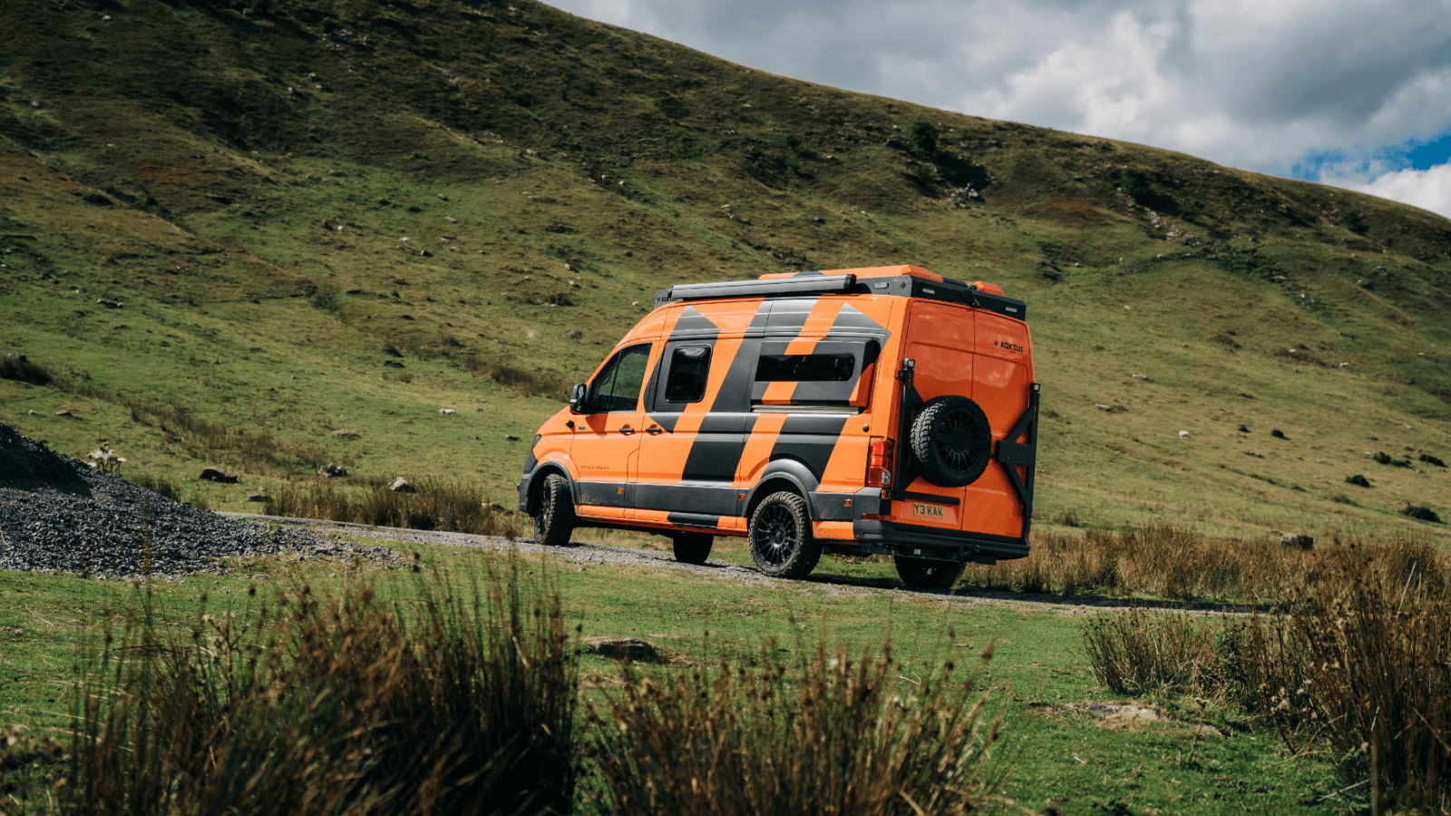 An orange van with reflective stripes parked on a gravel road, surrounded by green grassy hills under a brand design-inspired cloudy sky.