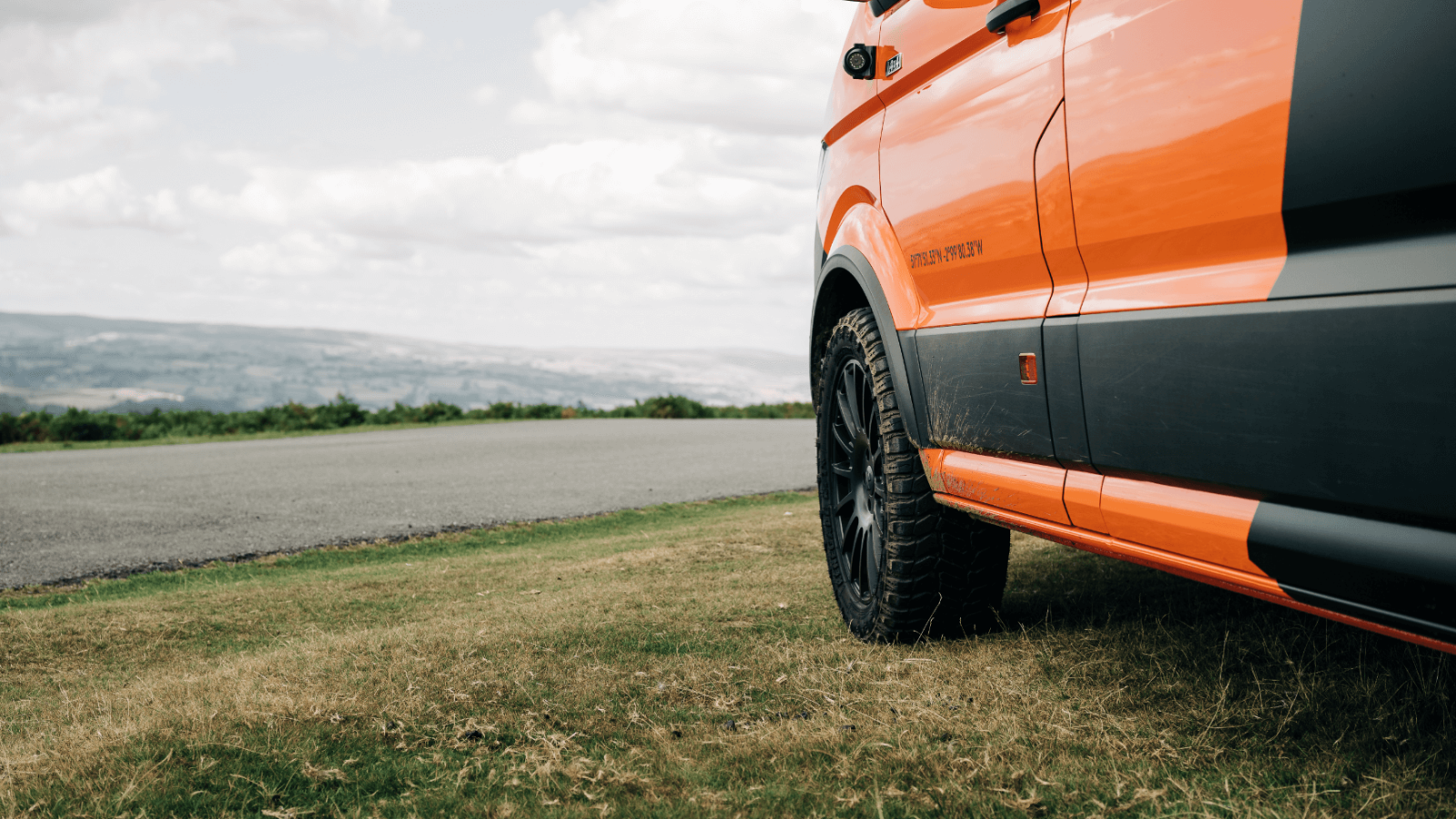 Close-up of a brand design bright orange van parked on the roadside, overlooking a scenic landscape with lush greenery and a cloudy sky in the background.