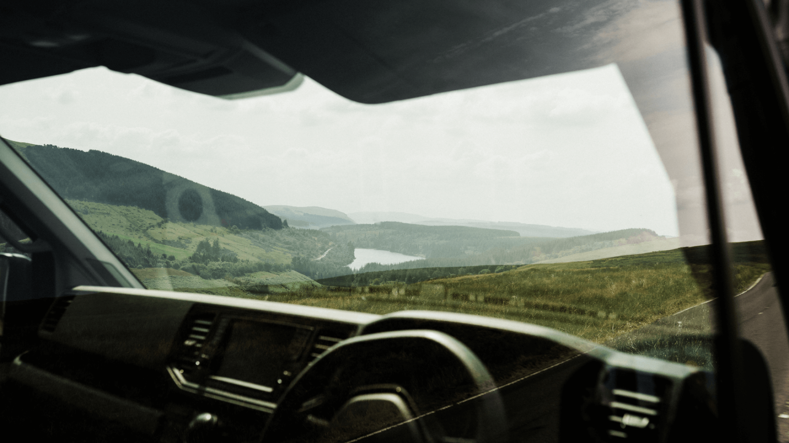 View from inside a car overlooking a scenic landscape featuring rolling hills and a winding river, with the dashboard displaying brand design visible in the foreground.
