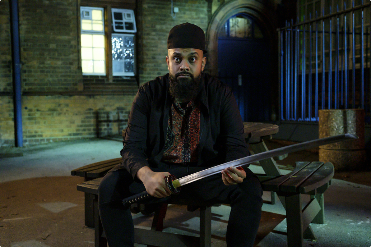 A bearded man wearing a black hat and suit sits on a bench at night, holding a long sword with a serious expression. street lights illuminate the scene.