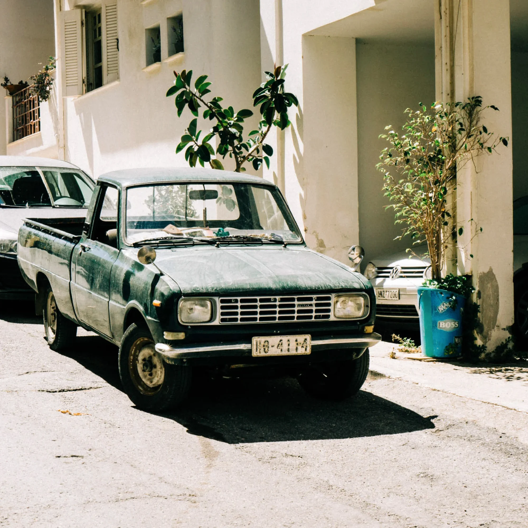 Old, dusty blue pickup truck parked on a sunny street beside a building, with lush green plants and a blue trash bin nearby. License plate visible. Ideal for a design agency UK photo shoot setting.