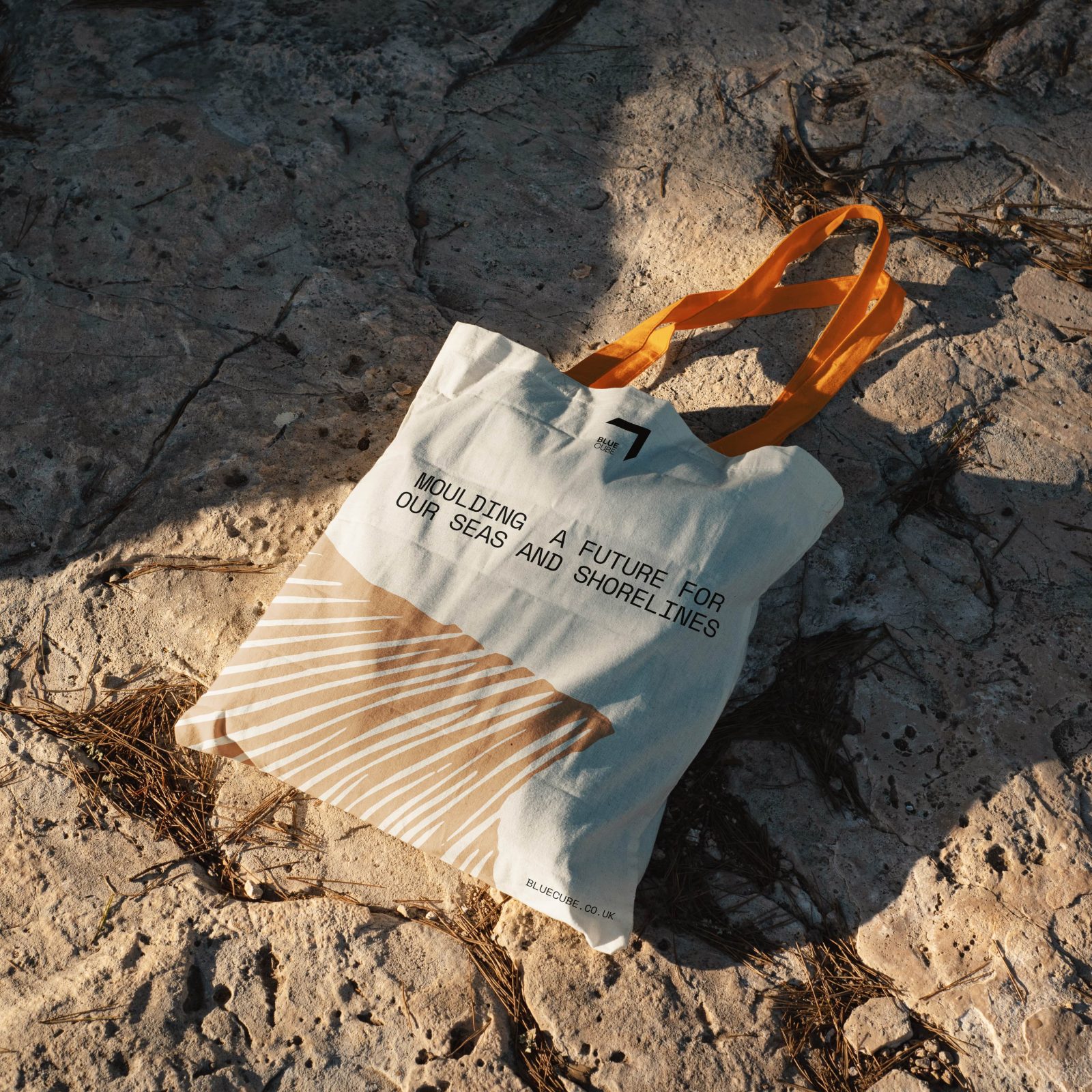 A canvas tote bag with orange handles and brand design about cleaning seas lies on a rocky surface under sunlight, casting a shadow.