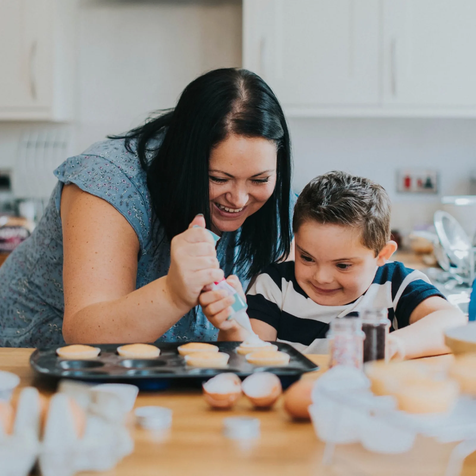 A woman and a young boy happily baking together in a kitchen, with the boy using a whisk on a bowl of batter as they both smile joyfully, evoking the feel of website design that is
