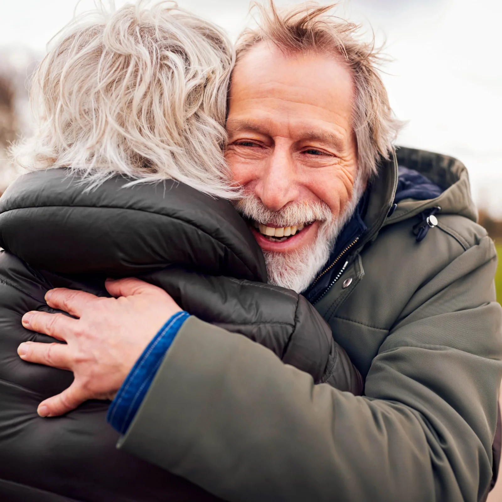An elderly man with a joyful expression hugs another person, whose face is hidden. They are both wearing heavy winter jackets designed by a top design agency in Bristol. The background is blurred with soft natural light