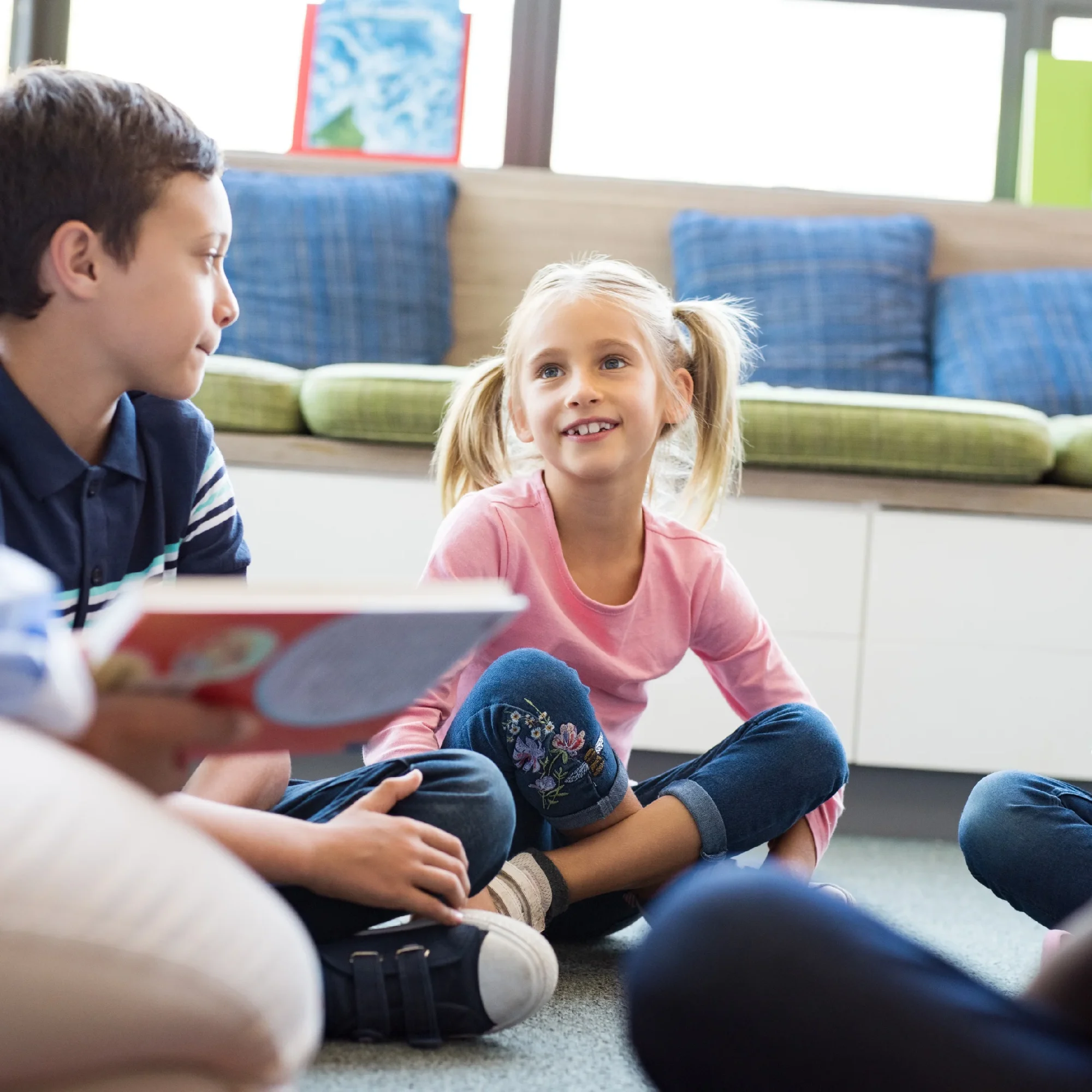 Two children sitting on the floor in a colorful classroom. A boy is reading a book to a girl who is smiling and listening attentively.