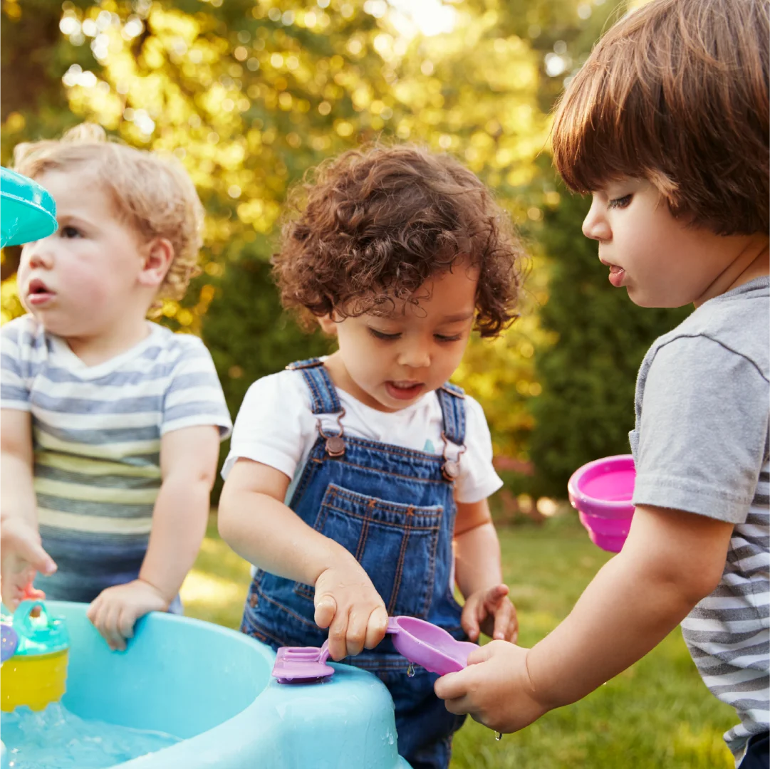 Three toddlers playing with water and toys in a backyard. One child in overalls pours water, while another watches, and the third plays nearby. They are surrounded by toys, including a new website development