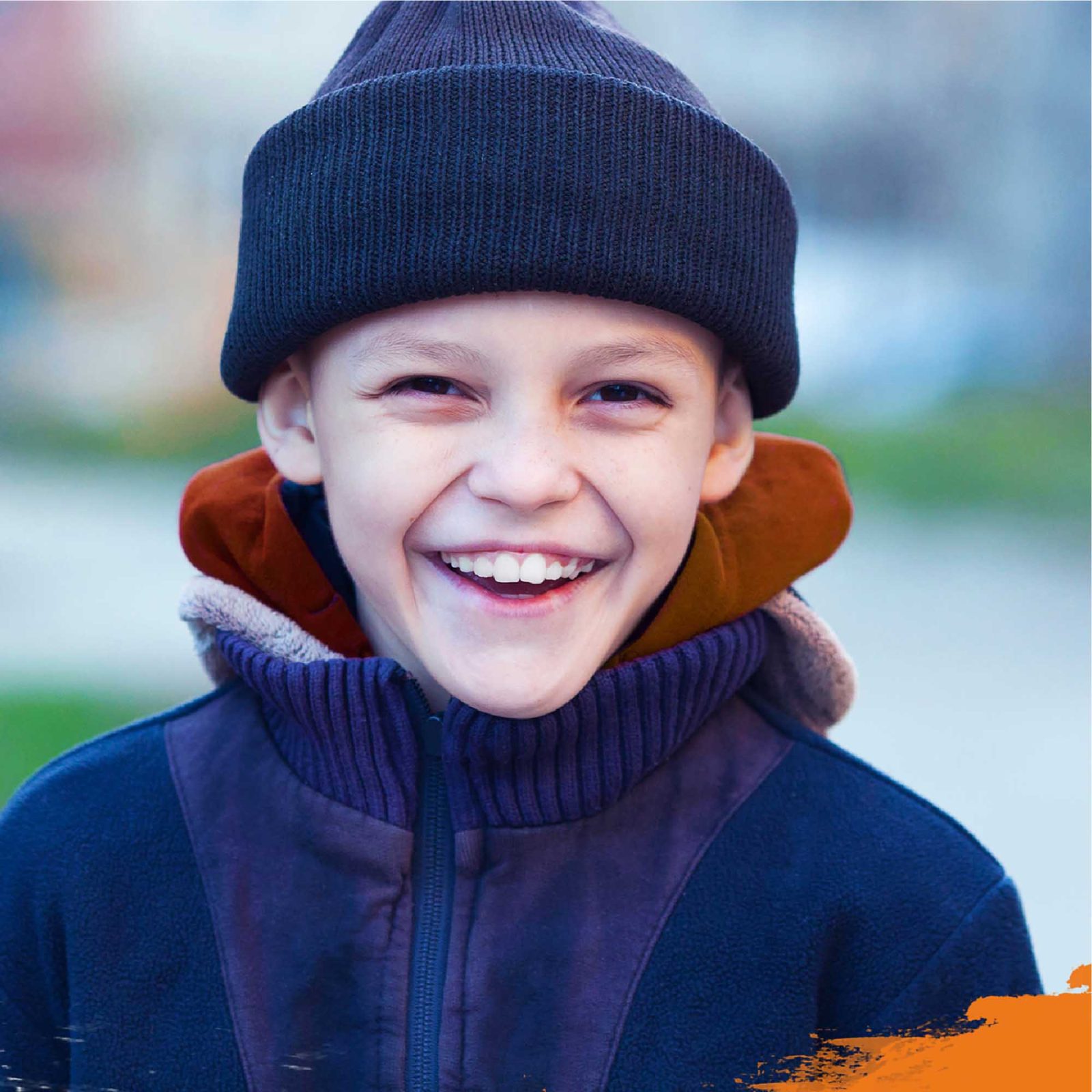A young boy with a joyful smile, wearing a black beanie and blue jacket designed by a renowned brand design agency, stands outdoors. His cheeks are rosy, and his eyes gleam with happiness