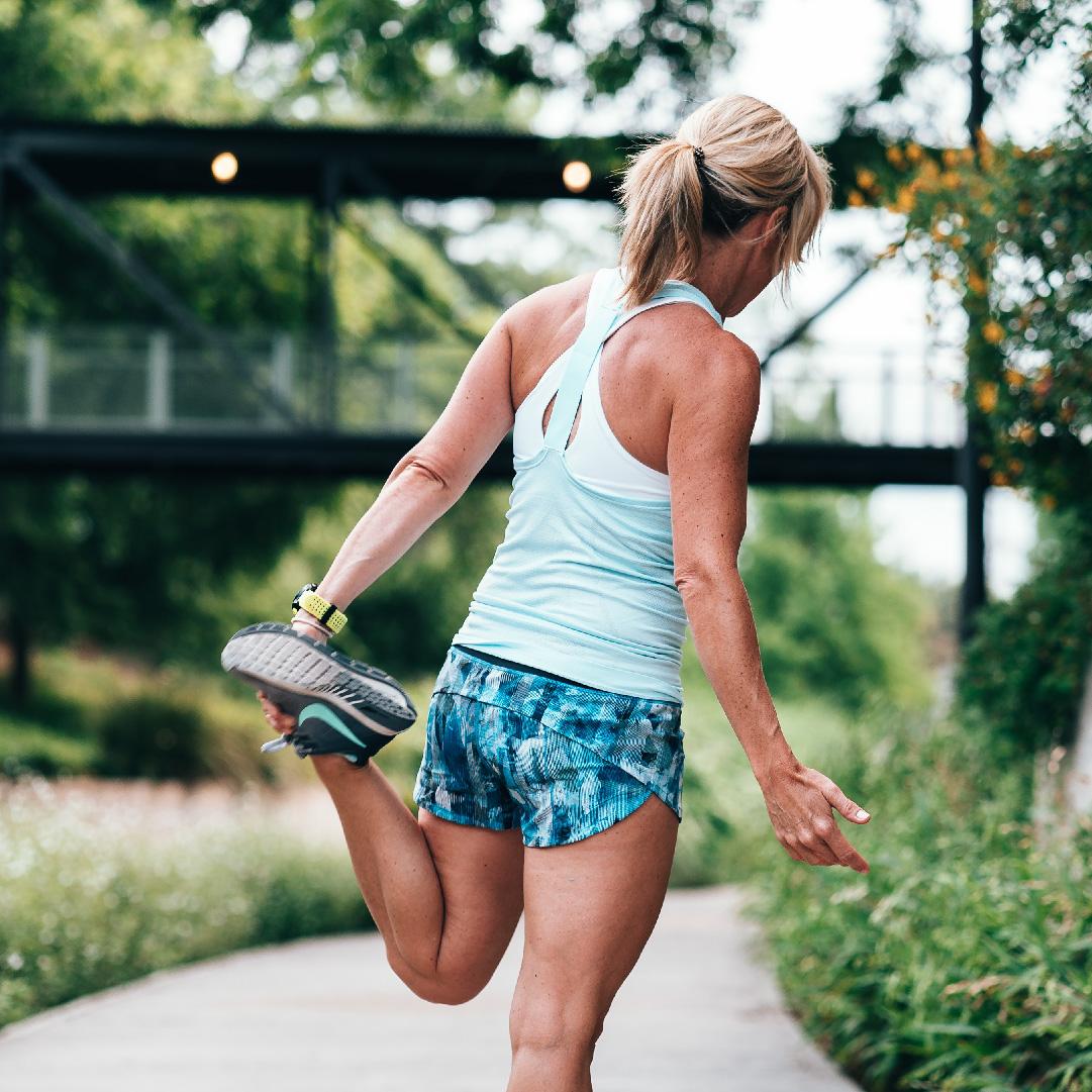 A woman in workout attire stretches by pulling her heel towards her buttocks, standing on a path with greenery and a bridge in the background, showcasing the influence of brand design on active lifestyle imagery.
