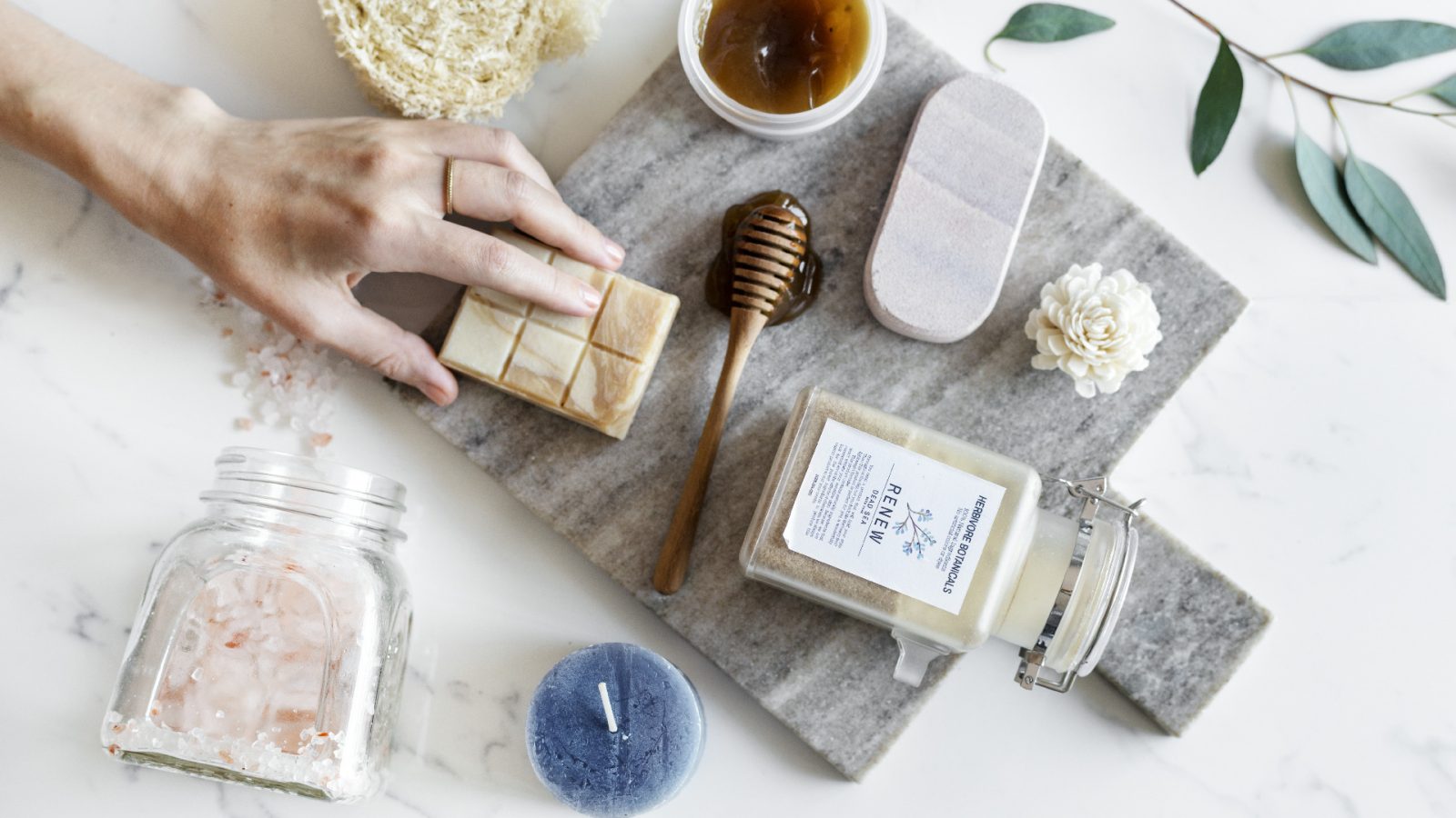 A person's hand reaching for a bar of soap on a marble countertop surrounded by various spa items including a honey dipper, candles, scrub, salt, and a small white flower designed for brand