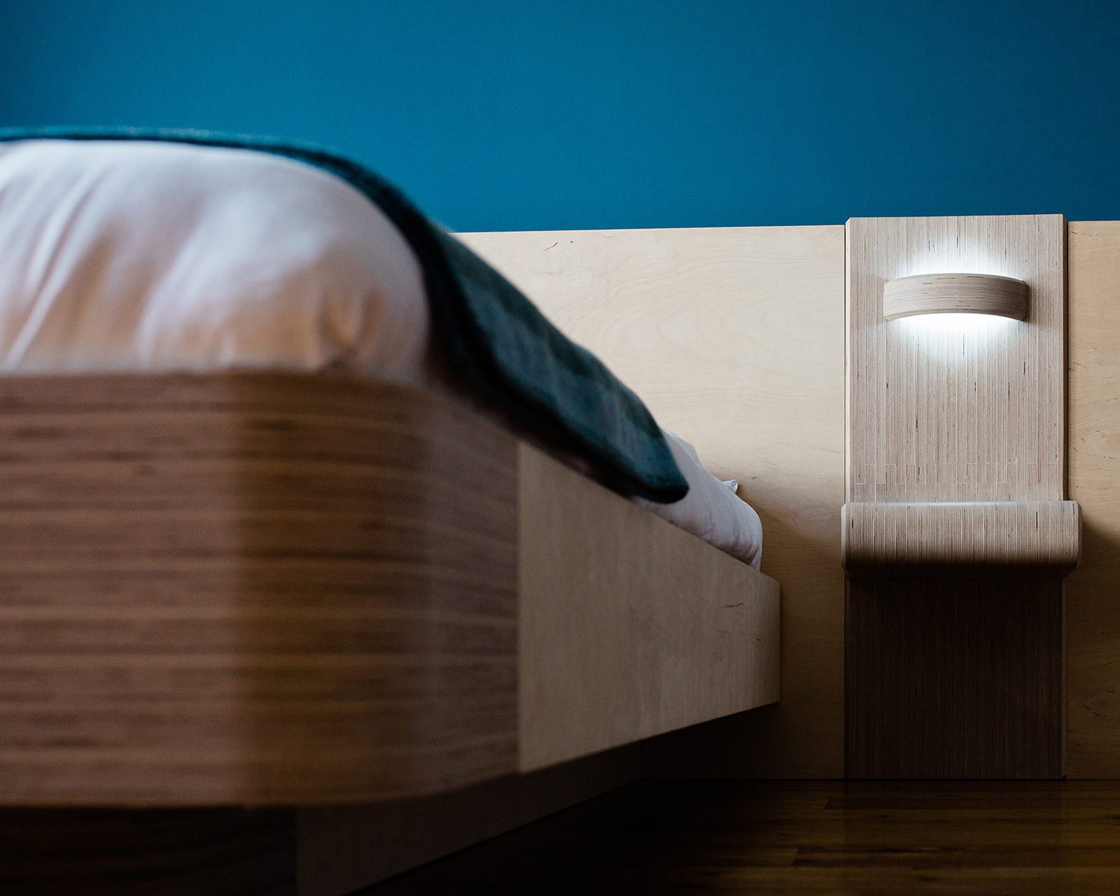 A modern wooden bed frame and attached bedside table in a room with blue walls and natural light illuminating the scene, crafted by a top Design Agency UK.