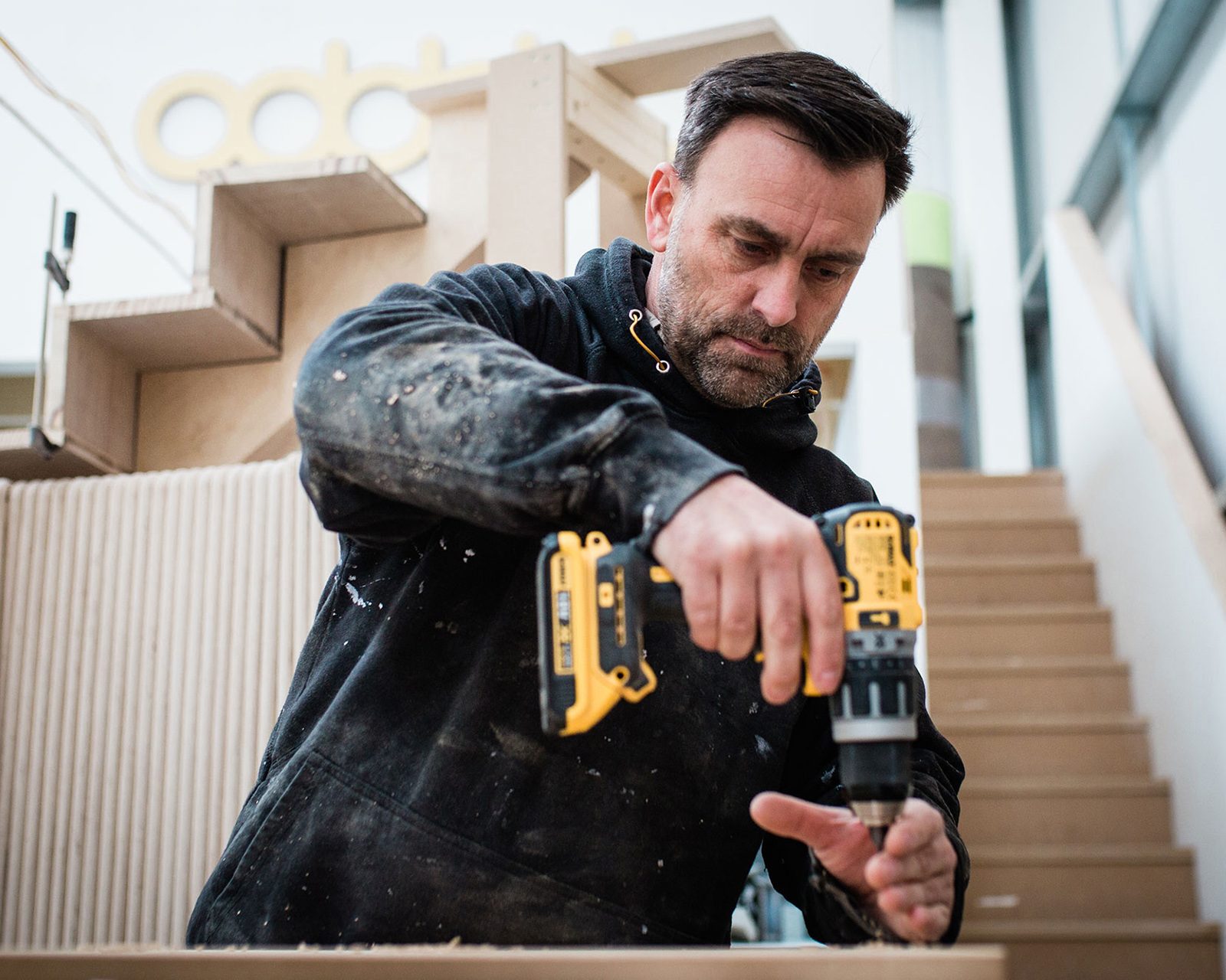 A focused man with a beard, wearing a dirty black jacket, uses a yellow power drill on a piece of wood at a construction site with brand design cardboard and wooden structures in the background.