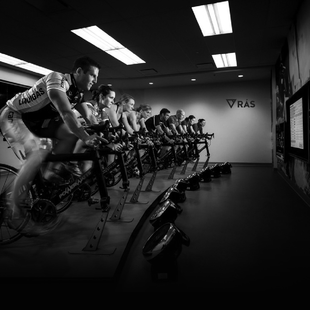 A black and white image of a group of cyclists, both men and women, in a row engaging intensely in a stationary bike training session indoors, possibly for a Website Design campaign.