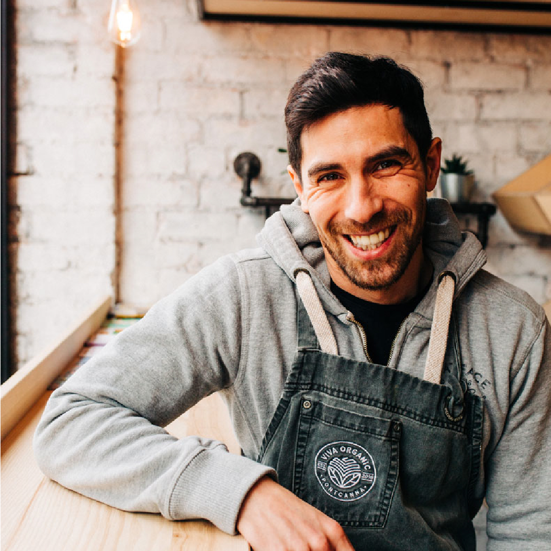 A smiling man wearing a gray hoodie and a black apron sits at a wooden table, with a blurred brick wall in the background at Brand Agency UK.