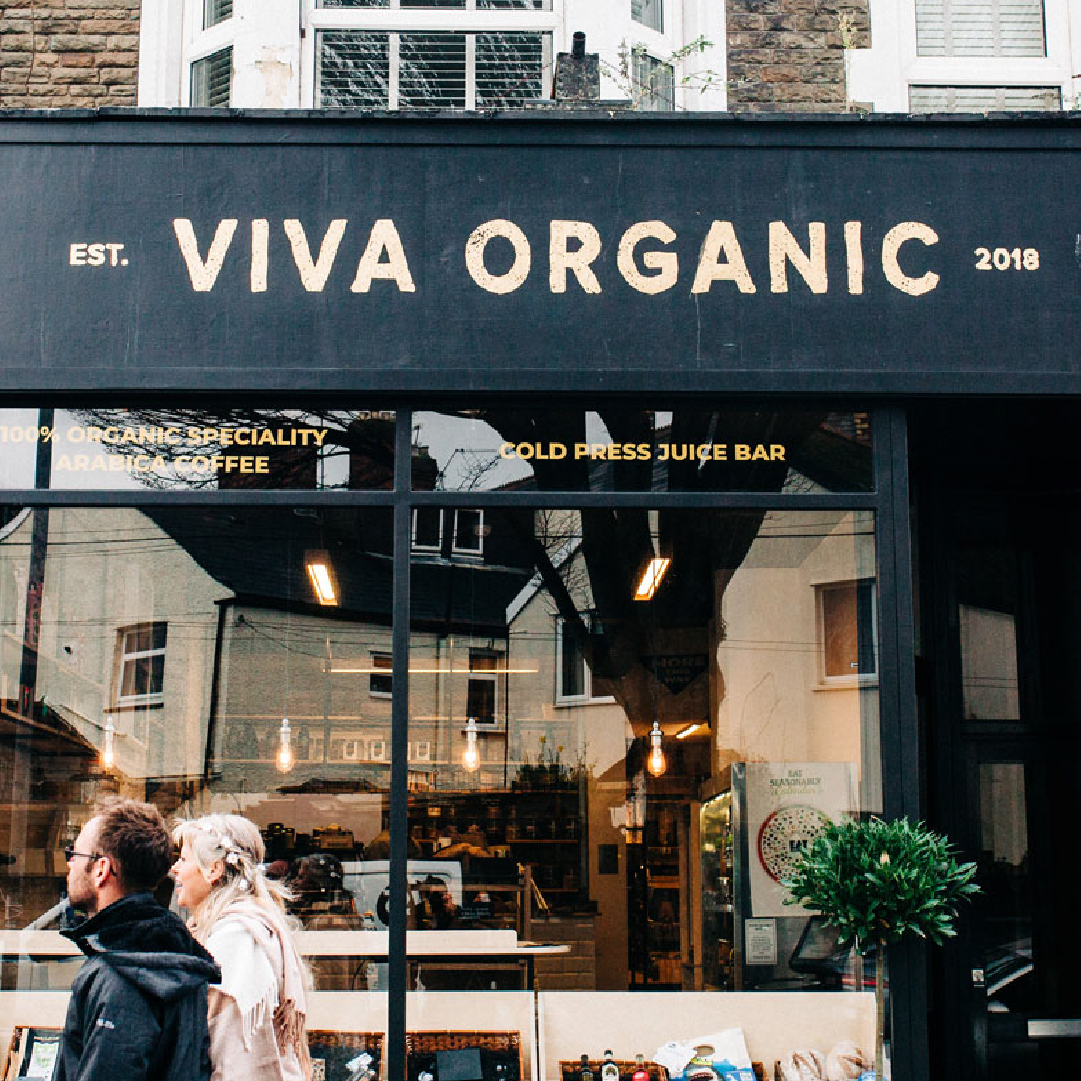 A couple walks by Viva Organic, an establishment with a black sign, founded in 2018, advertising organic specialties and a cold press juice bar. The shopfront has large windows displaying indoor plants