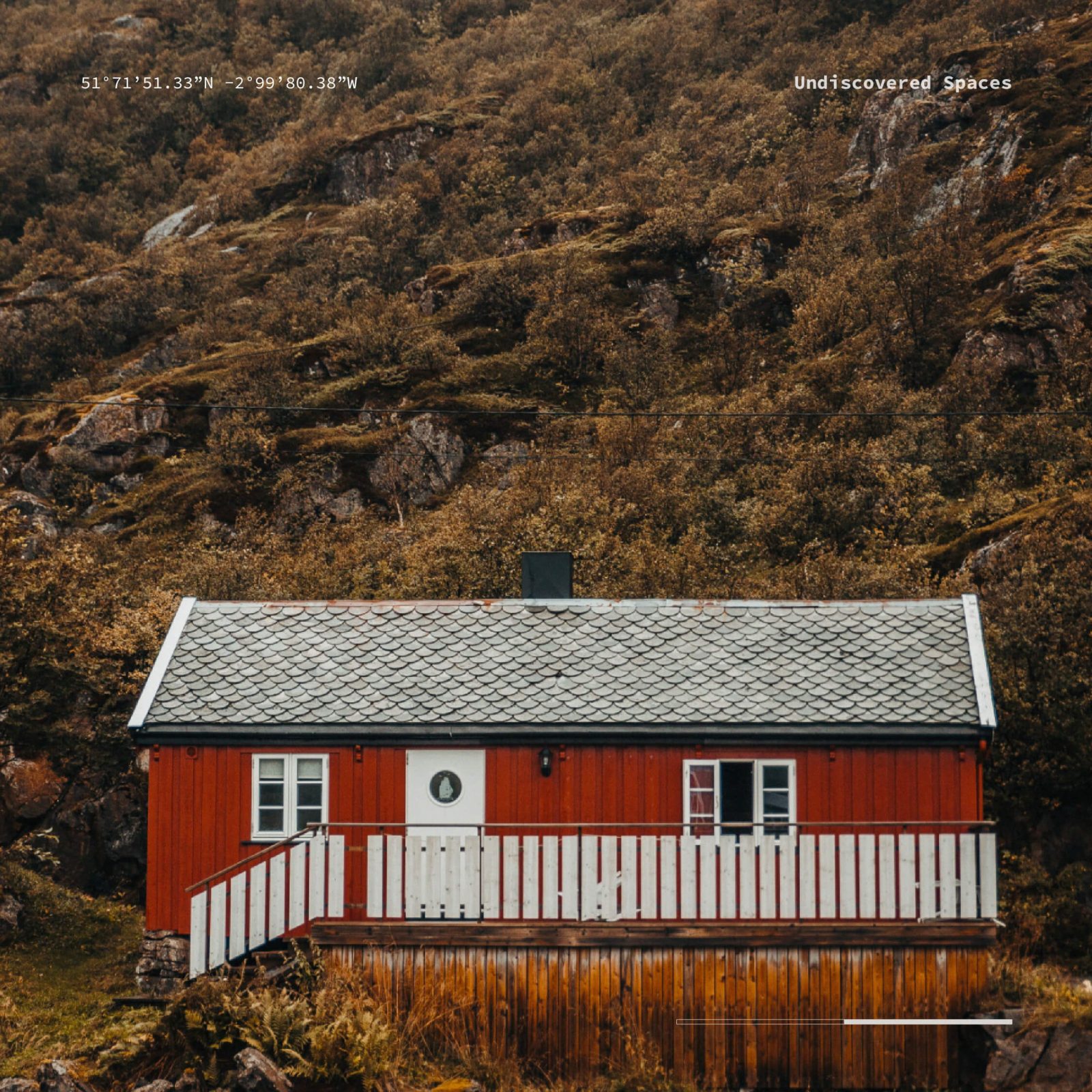 A small red cabin with a white trim and a black roof is nestled against a rugged, rocky hillside covered with sparse green vegetation. Mist hovers around the landscape, enhancing its natural appeal much like