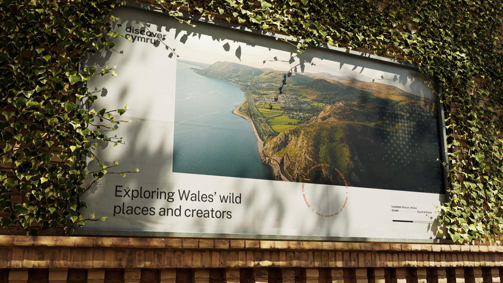 A large outdoor advertisement on a brick wall, partially covered with ivy, showcasing an aerial photo of a coastal landscape with the caption 