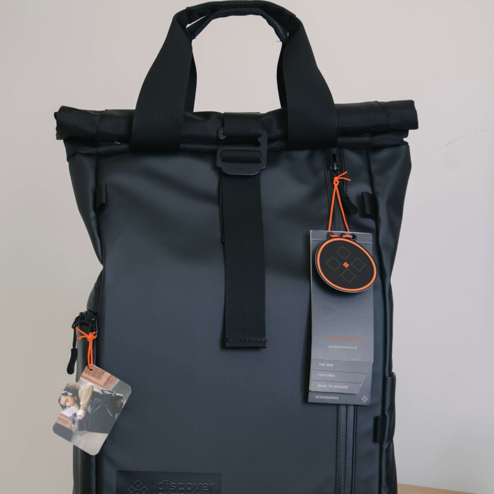 A gray roll-top backpack with black straps, featuring attached tags and a small compass, branded by a leading Brand Agency UK, standing upright against a light beige background.