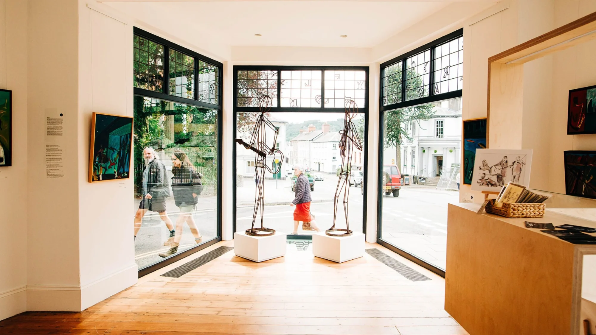 Interior of an art gallery with large windows and doors; sculptures on pedestals near the entrance. People are visible walking outside. Bright and welcoming ambiance, reminiscent of a design agency in Bristol.