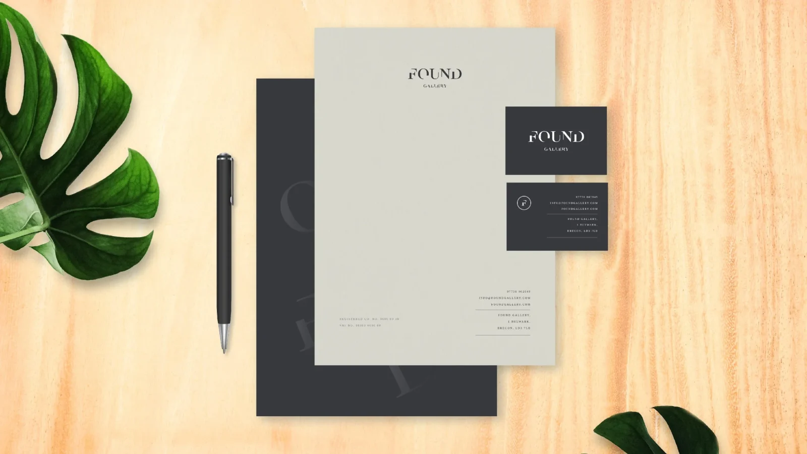 A neatly arranged stationery set on a light wooden surface, showcasing a gray business folder, brand design business cards, and a pen, accompanied by a green leafy plant.