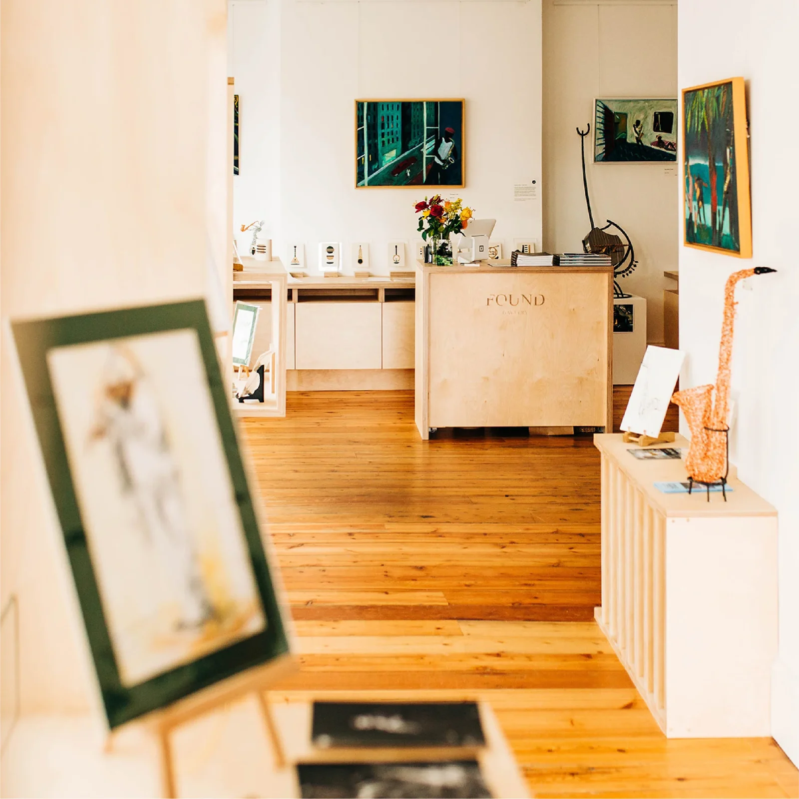 A warm, welcoming art gallery with wooden floors showcasing various artworks and sculptures. A reception desk labeled 