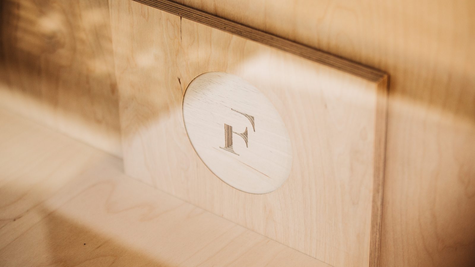A wooden panel with an engraved circular detail featuring the letter “f” in a serif typeface, casting a soft shadow on the surrounding wood surface, ideal for brand strategy visuals.