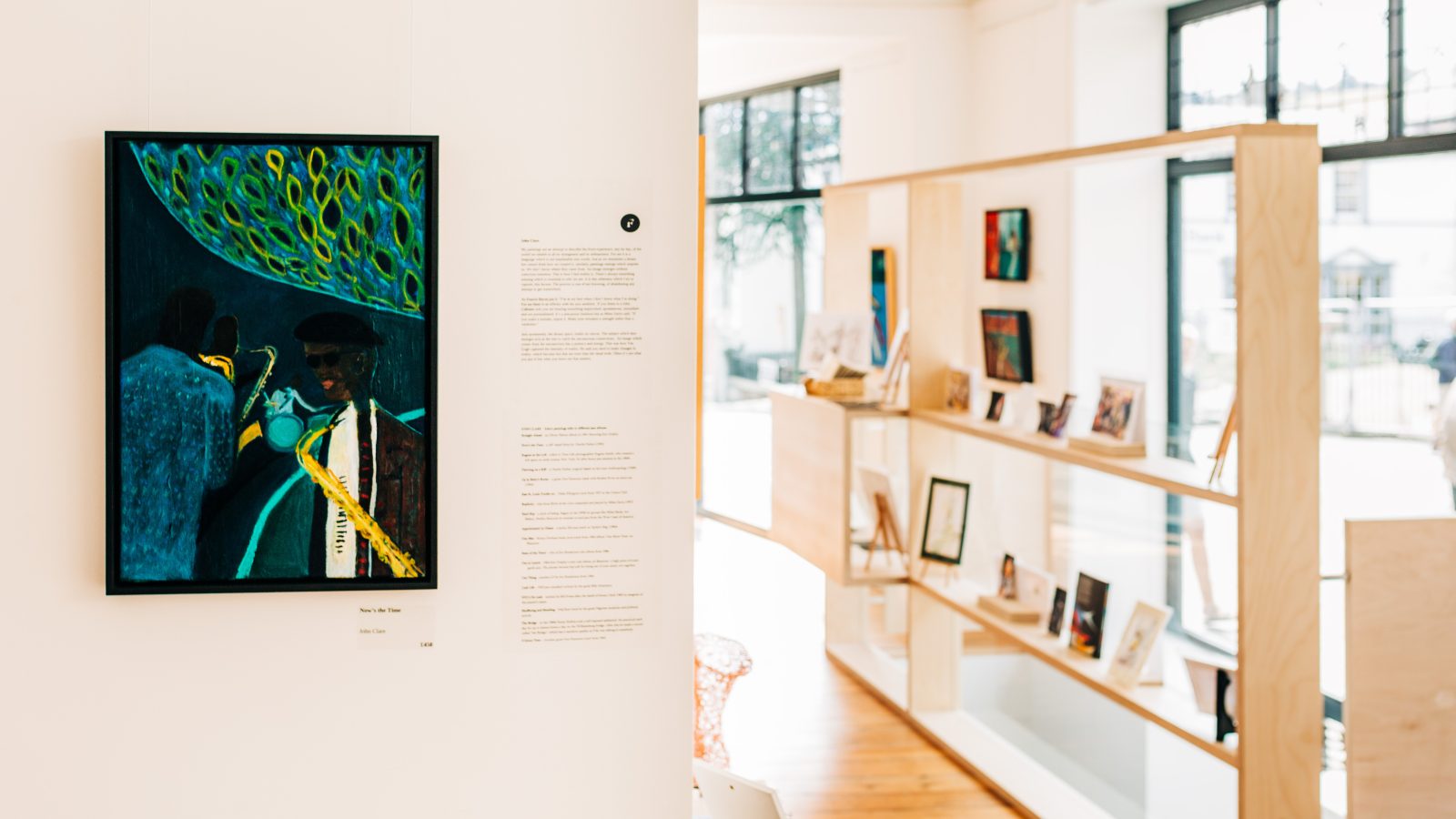 Art gallery interior featuring a vibrant painting on the left wall, with a visitor reading an informational plaque beside it. Shelves with framed artworks are visible across the room near a bright window, subtly showcasing the