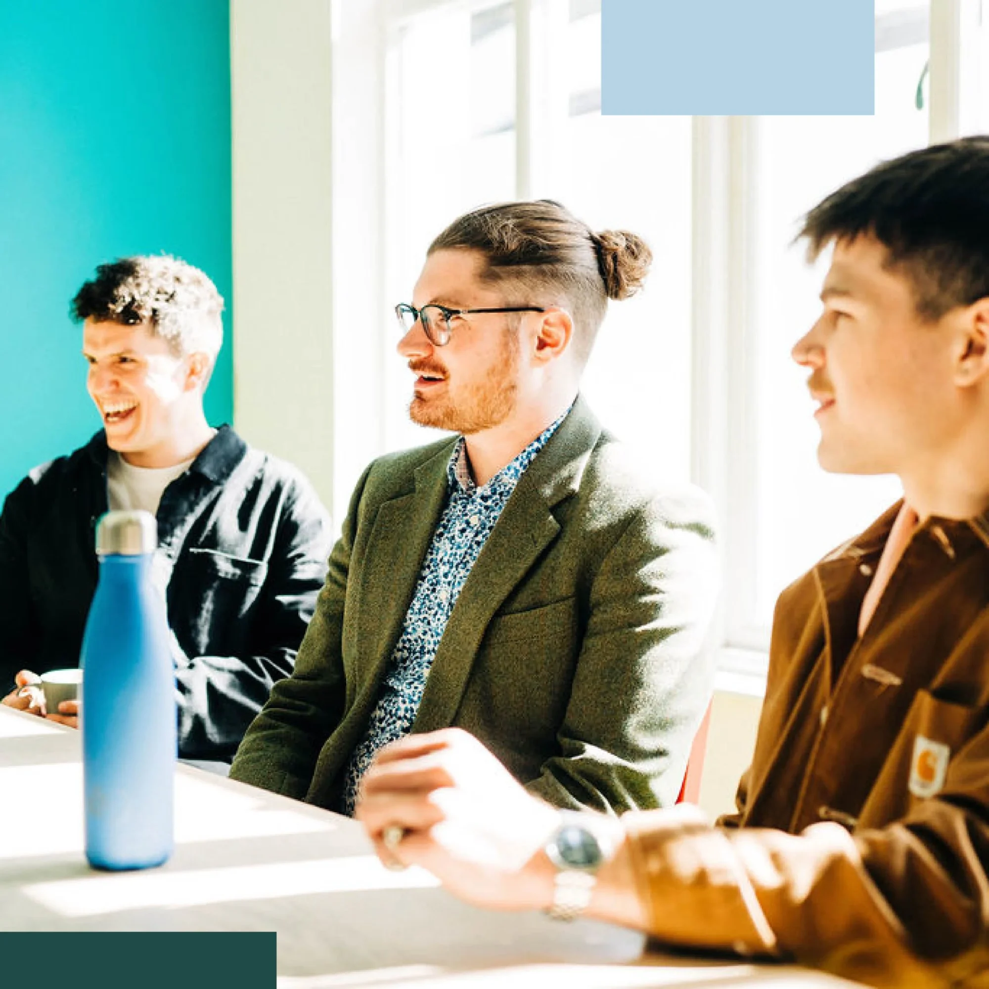 Three men of various appearances sitting at a table engaged in a discussion in a brightly lit room with large windows. a blue water bottle is visible on the table.