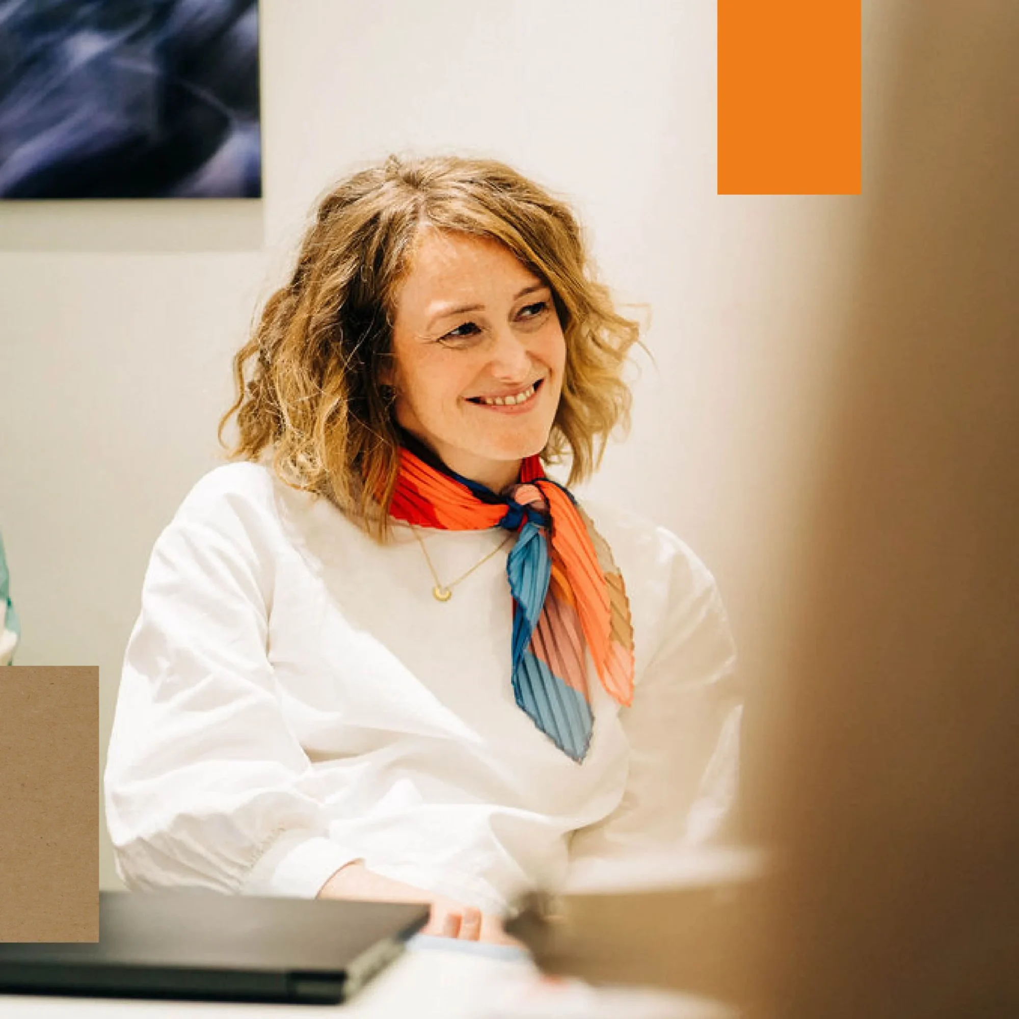 A woman with curly hair, wearing a white shirt and a colorful scarf, smiles while looking at a colleague in an office setting.