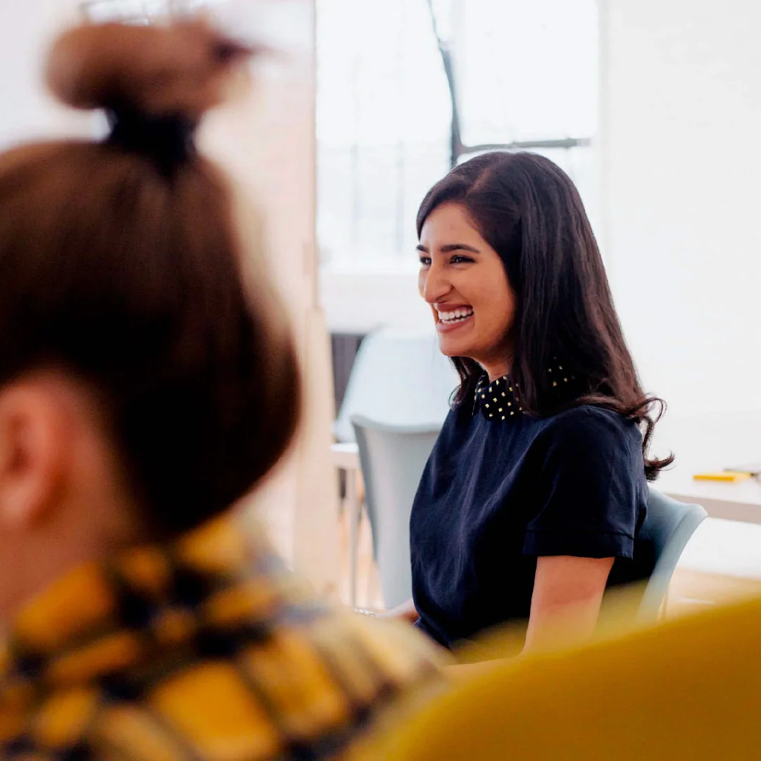 A joyful woman with long dark hair tied up in a bun, wearing a navy-blue top and decorated collar, smiling while sitting in a brightly lit room, epitomizes the brand design of our Brand
