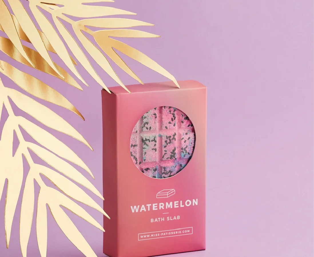 A pink watermelon-scented bath slab box with a transparent window showing the product inside, designed by a top Design Agency Bristol, set against a pink background with a decorative metallic gold palm leaf.