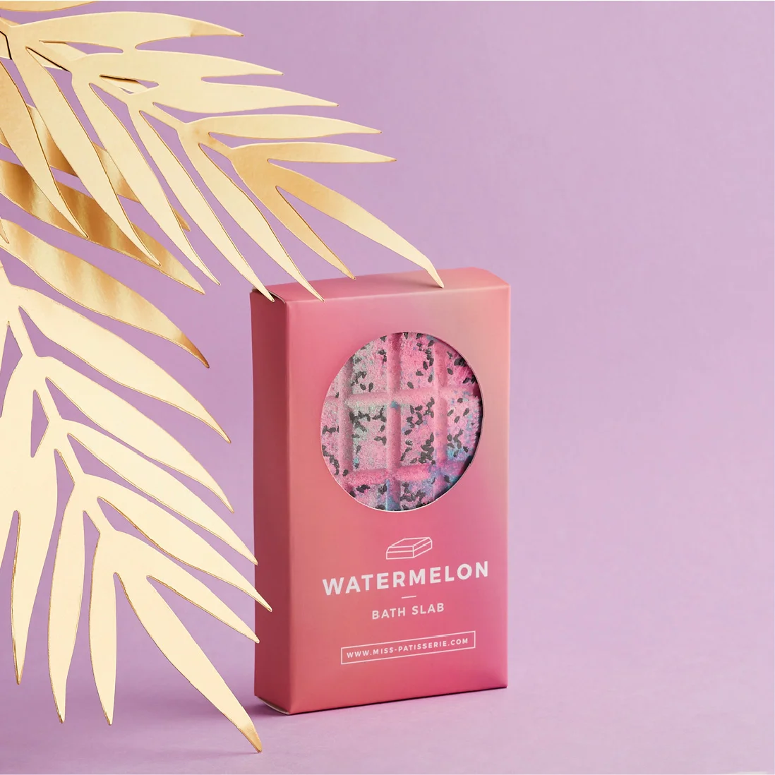 A pink watermelon-scented bath slab box with a transparent window showing the product inside, designed by a top Design Agency Bristol, set against a pink background with a decorative metallic gold palm leaf.
