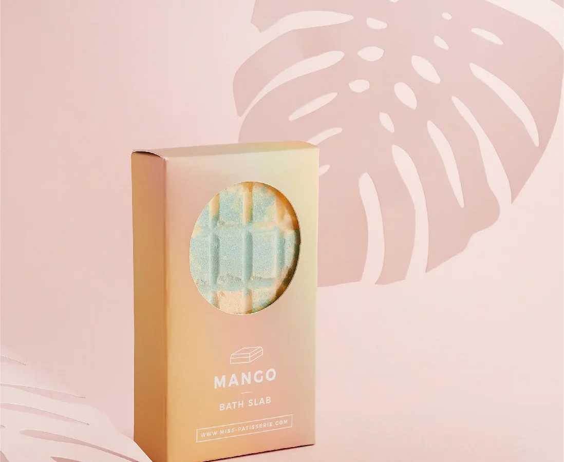 A mango-scented bath slab in a gold packaging with a circular window revealing the product, set against a soft pink background with a white monstera leaf silhouette, designed by a top design agency