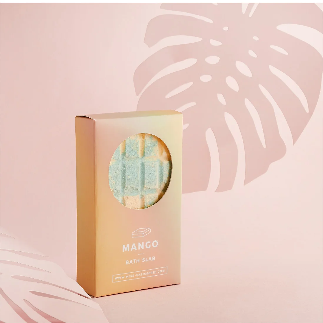 A mango-scented bath slab in a gold packaging with a circular window revealing the product, set against a soft pink background with a white monstera leaf silhouette, designed by a top design agency