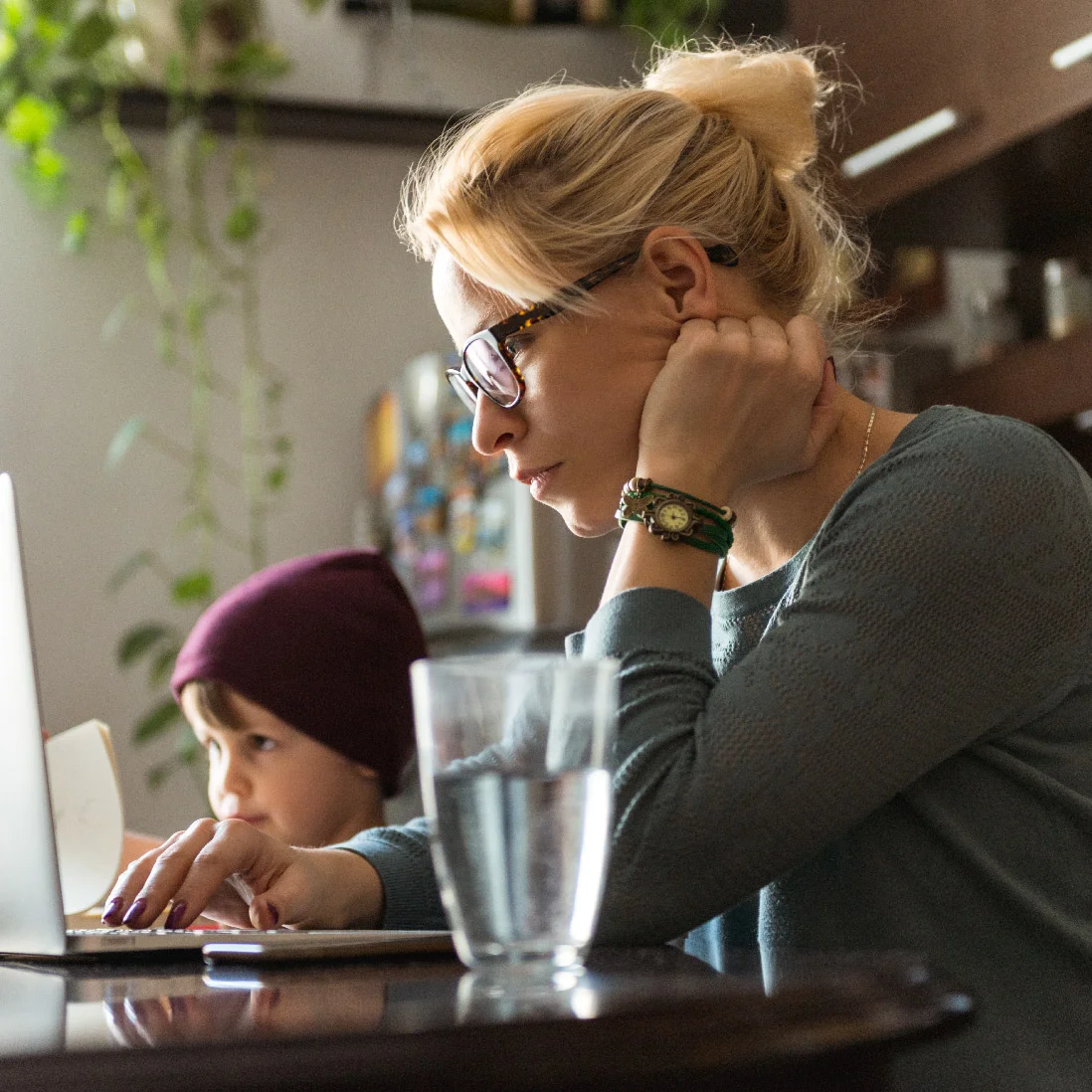 A woman with glasses and a bun, wearing a green sweater, uses a laptop intently to work on website design while a young child in a maroon beanie looks on, both seated at a