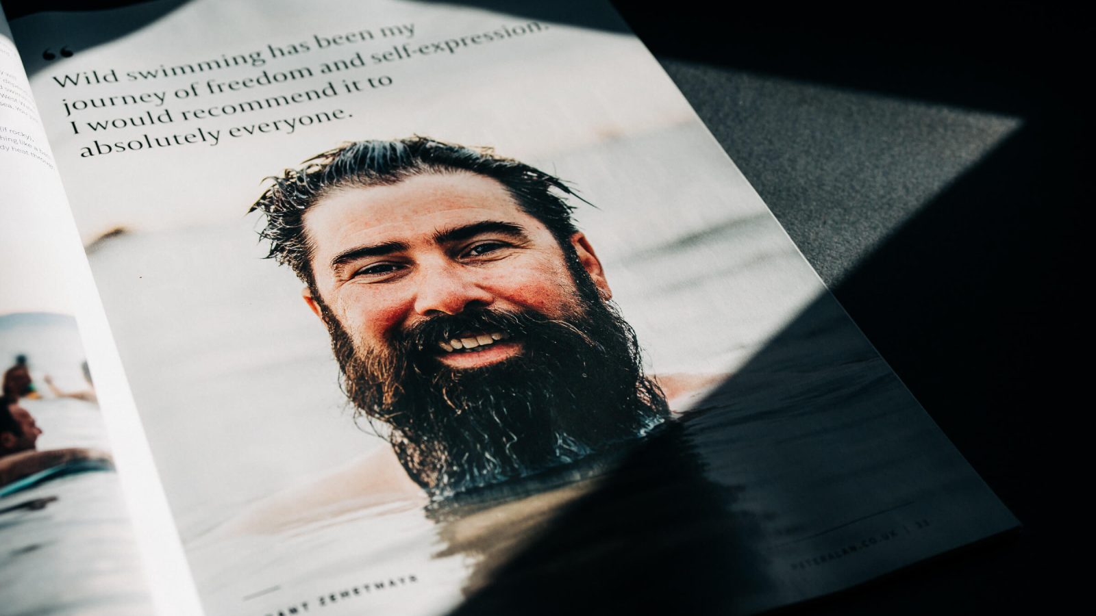 A magazine lies open to a page featuring a portrait of a smiling bearded man in water, with a quote about enjoying wild swimming, partially cast in shadow by the Design Agency UK.