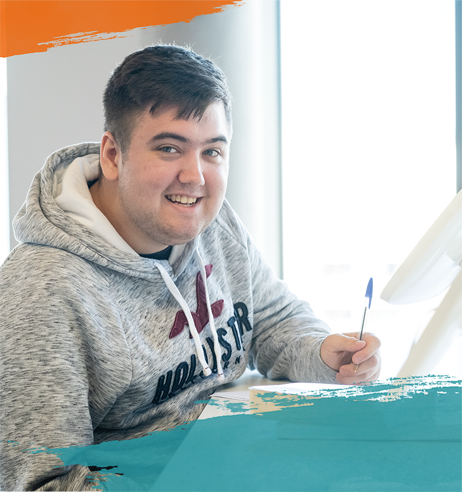 A young man wearing a Hollister hoodie smiles while holding a pen, sitting at a desk with papers in a brightly lit room of a Web Agency in Bristol.