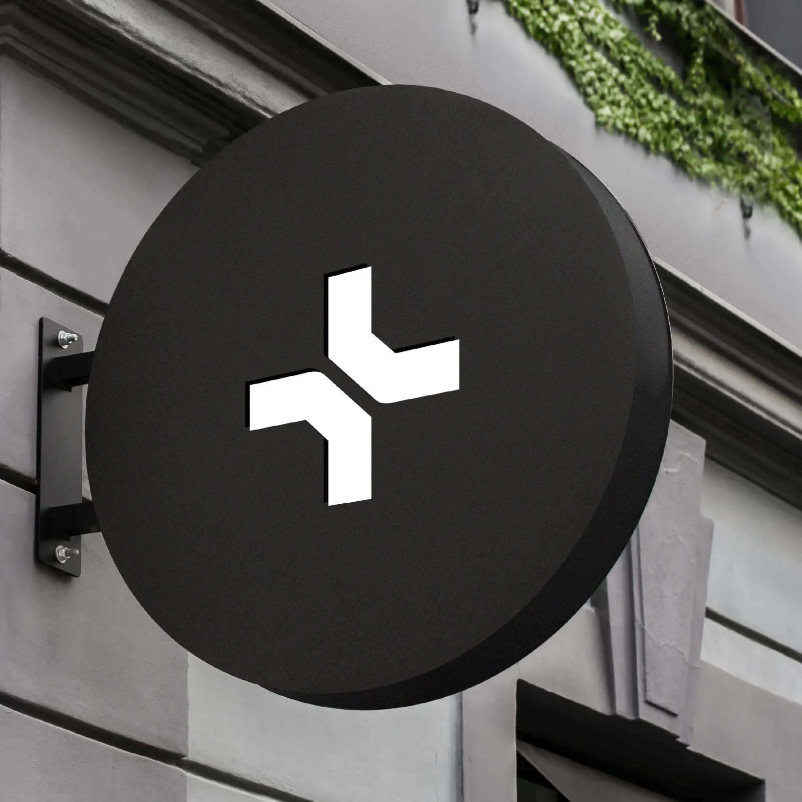 A circular black sign with a white arrow pointing left and branching to the upper right, designed by a design agency in Wales, mounted on a gray building facade next to green foliage.