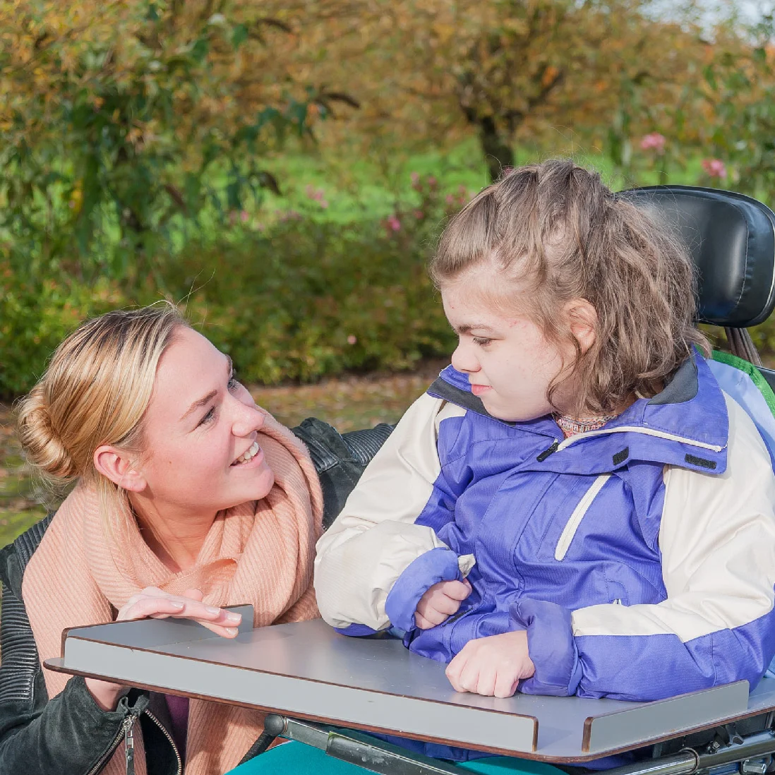 A smiling woman leans towards a young girl in a wheelchair, both enjoying a sunny day outdoors surrounded by autumn foliage. The girl, wearing a purple jacket, looks at the woman affectionately as they discuss