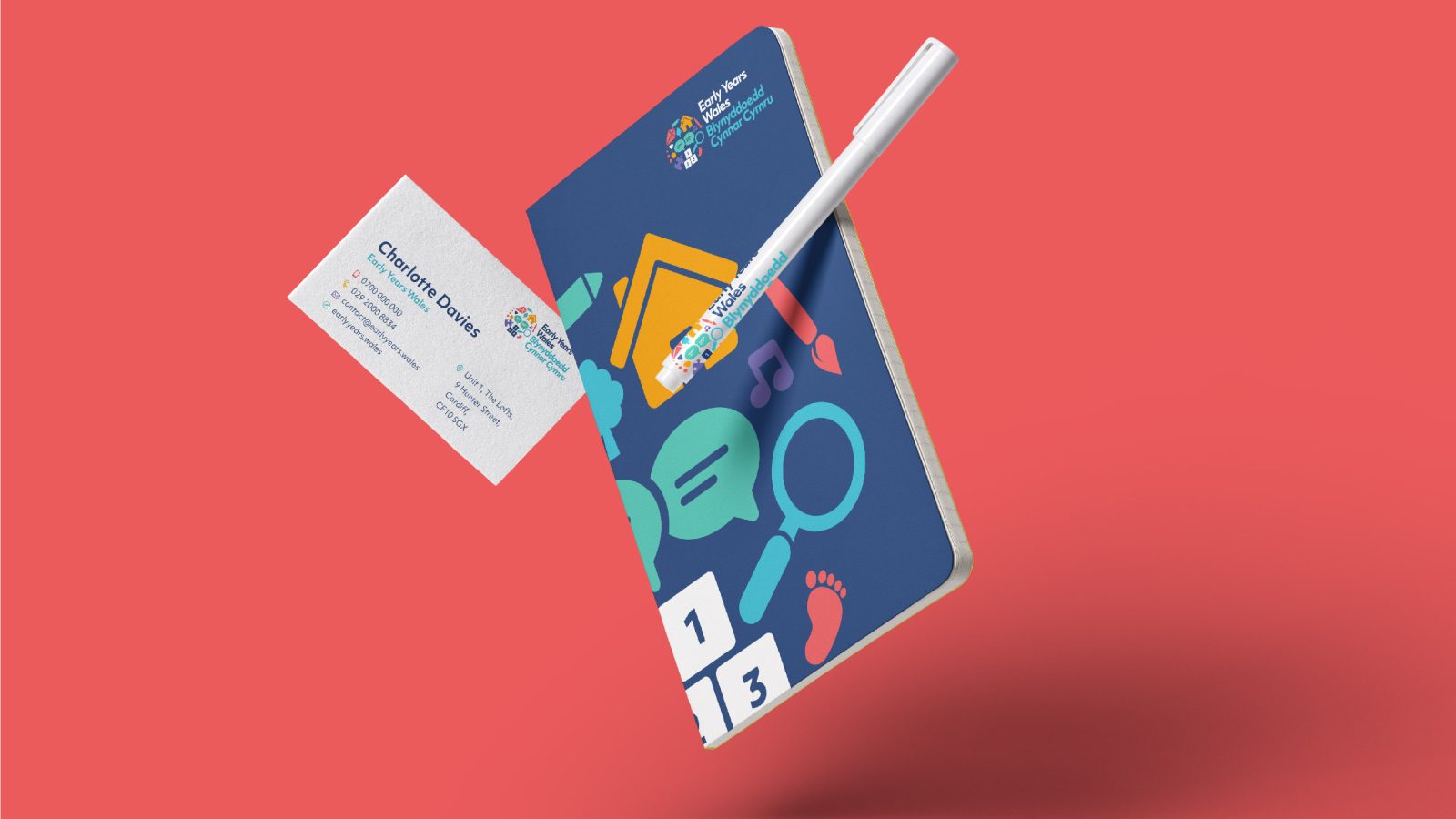 A realistic graphic of a planner, pen, and business card from a Design Agency UK floating against a vivid coral background. The planner displays a vibrant design with icons related to scheduling and tasks.