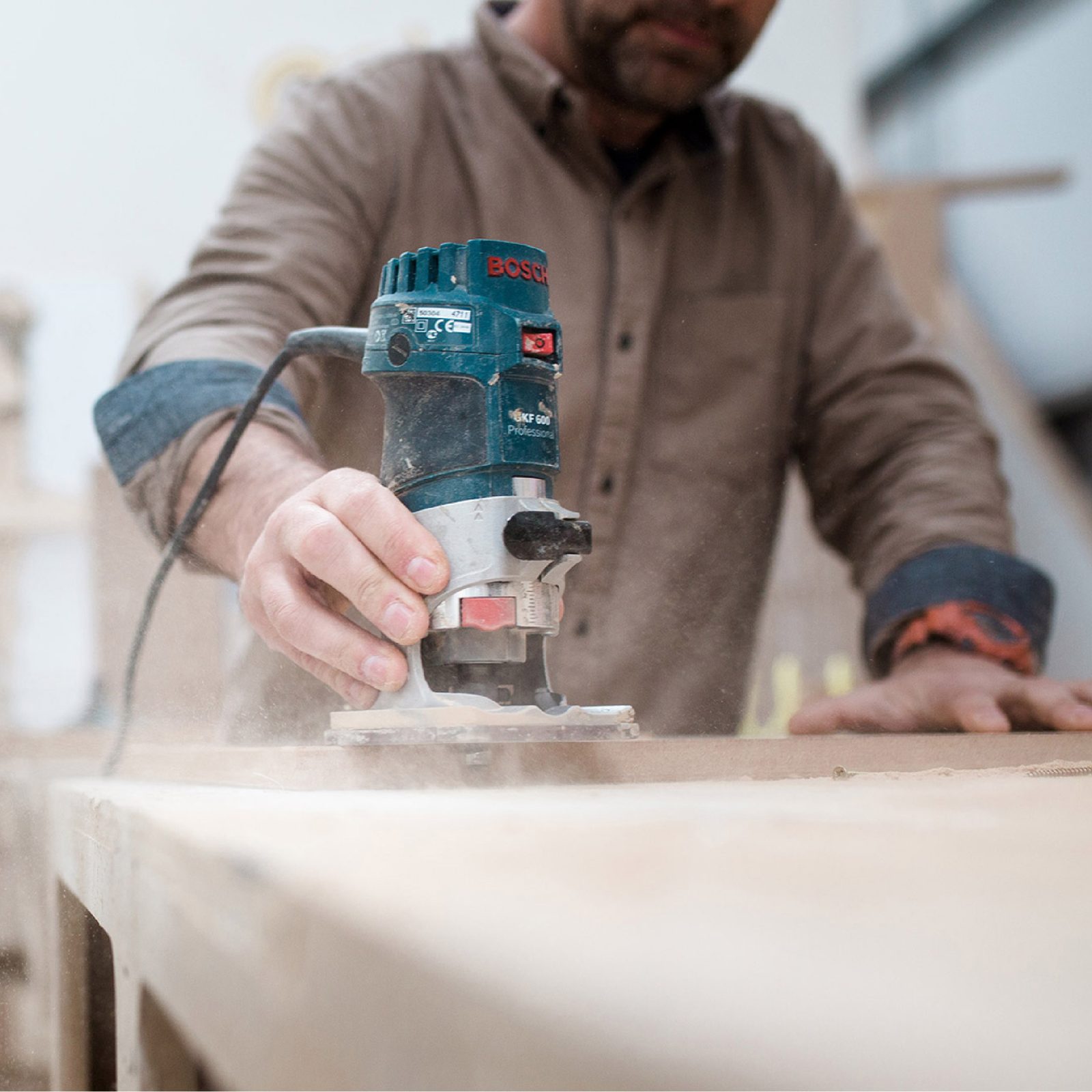 A man using a blue Bosch power tool to sand a wooden board in a workshop, with sawdust visible in the air. Focus on the hands and the tool.