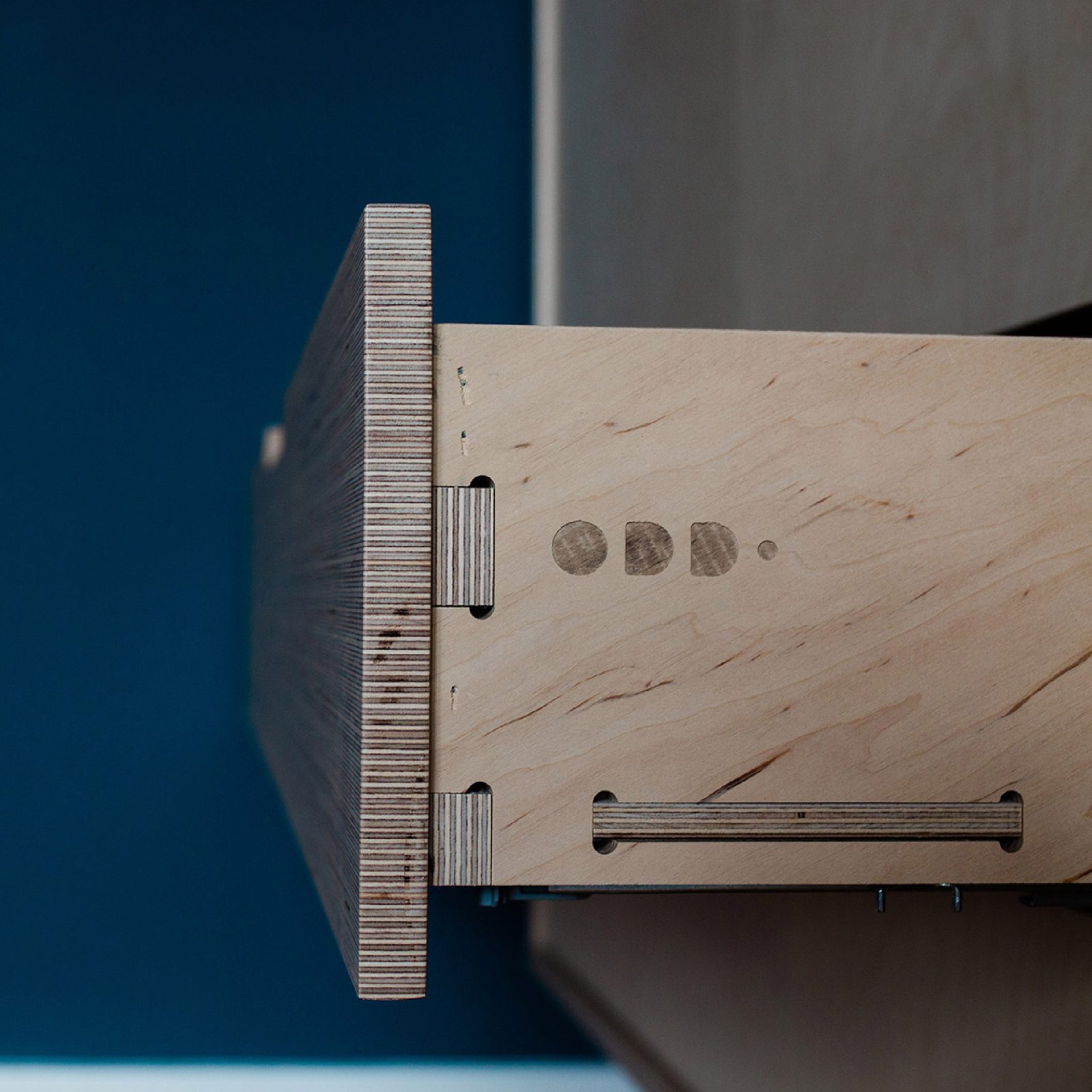 Close-up of a wooden drawer partially pulled out, showing detailed finger joints and brand design metal handle elements against a blue background.