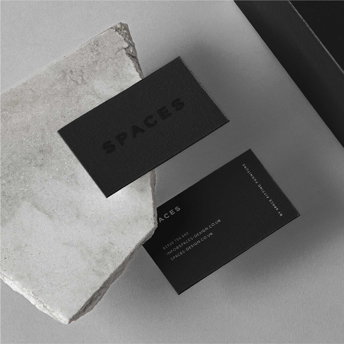 Two elegant black business cards with white text, one on a textured stone and the other partially obscured by it, placed on a smooth gray surface for a Brand Agency UK.