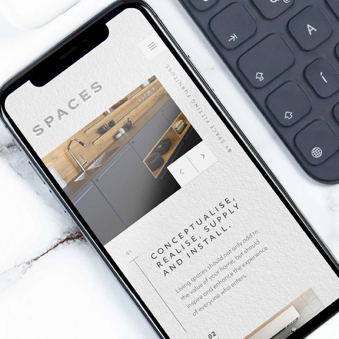 A smartphone displaying a kitchen design website, resting on a white marble surface next to a black keyboard. The phone screen features modern kitchen interiors by Design Agency UK.