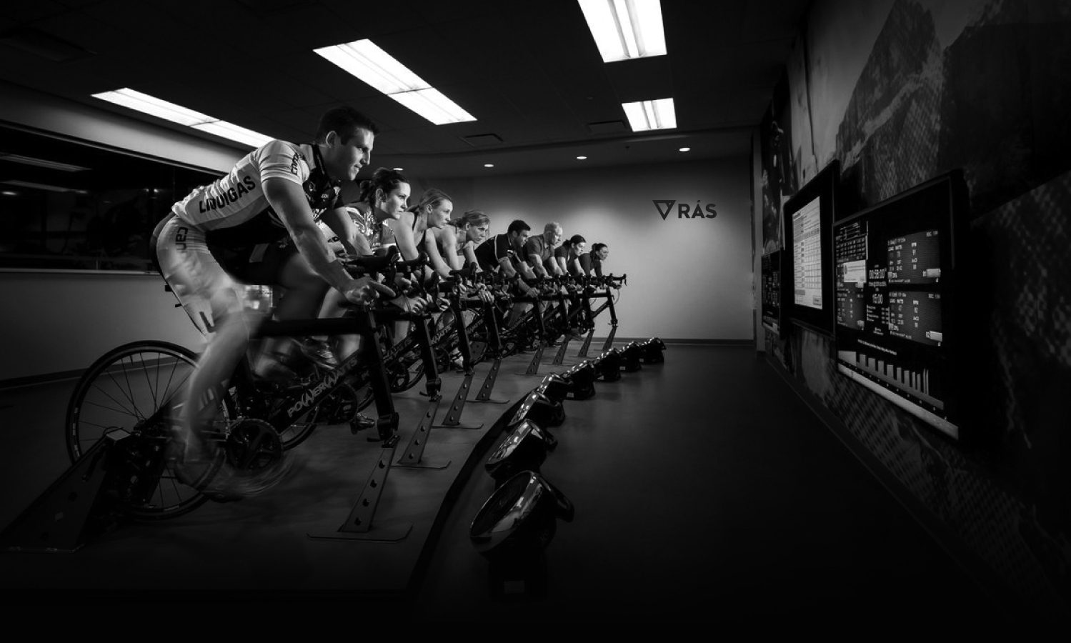 A group of cyclists training indoors on stationary bikes in a dimly lit room, focused intently on their workout, with a monitor displaying brand design performance metrics on the right.