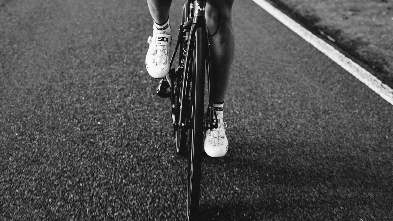 Black and white image focusing on the lower half of a cyclist pedaling on a road, showing details of the bicycle and brand design cycling shoes.
