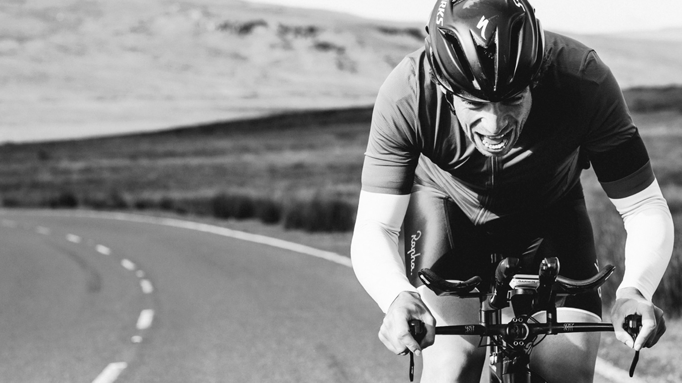 A cyclist in a helmet and sports attire aggressively pedals down a winding road through open, hilly terrain, in a high-intensity black and white scene designed by a Design Agency UK.