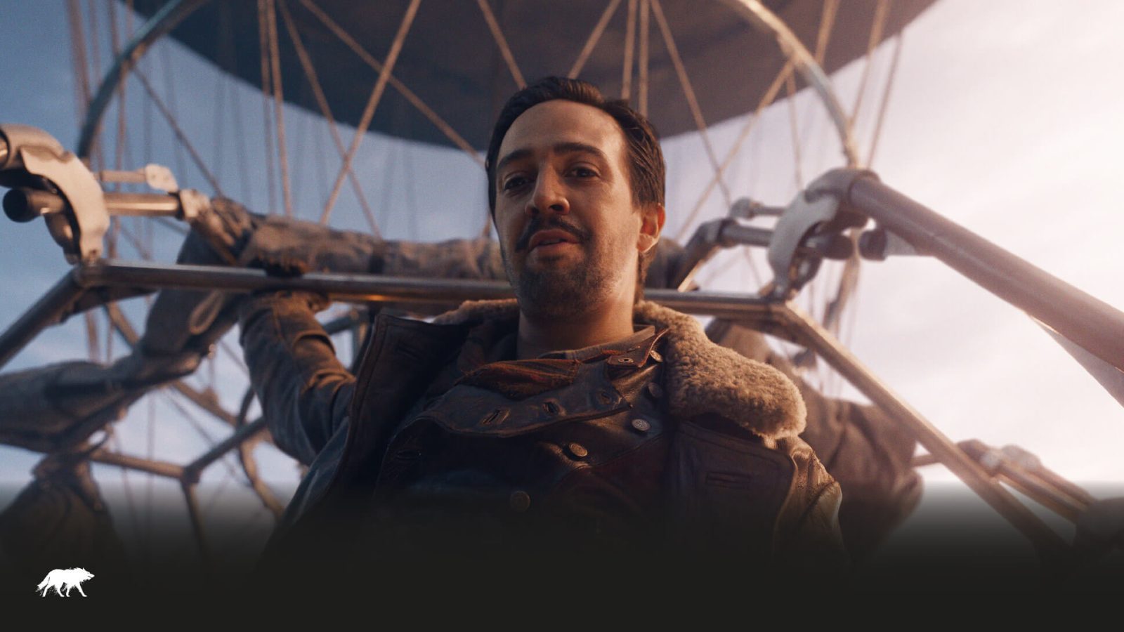 A man with a light beard wearing a leather jacket stands in front of a large metallic structure, designed by a prominent graphic design agency, which appears to be part of a futuristic ferris wheel, under
