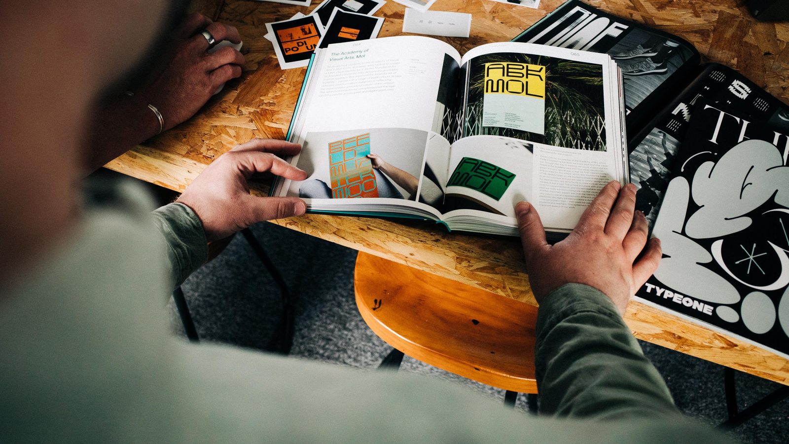 A person in a green jacket reading a design magazine opened to a page about typography at a brand design agency. Various print materials are scattered across a wooden desk.